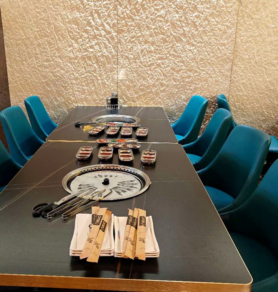 Grill table set for a group at Ombu Grill Orem, with banchan, tongs and teal chairs